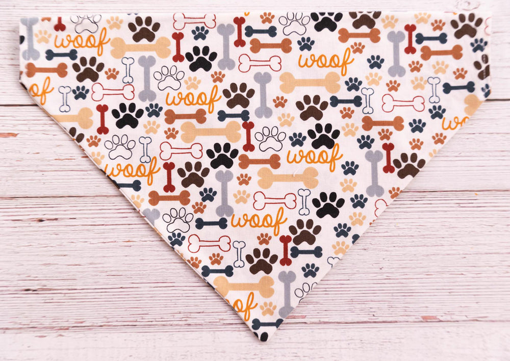 cream colored dog bandana with grey, black, brown, tan dog bones, paw prints and the word WOOF.